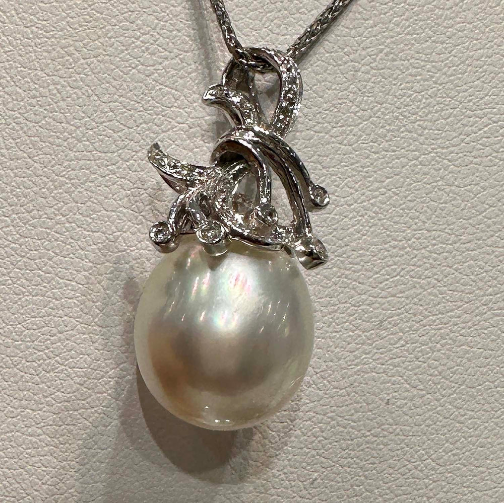 A close-up image of a pendant featuring a large South Sea pearl, suspended from an ornate silver setting adorned with small gemstones. The setting is designed with elegant, swirling motifs that add a decorative flourish around the pearl, which displays a lustrous sheen with subtle iridescent colors. The pendant hangs from a fine silver chain.