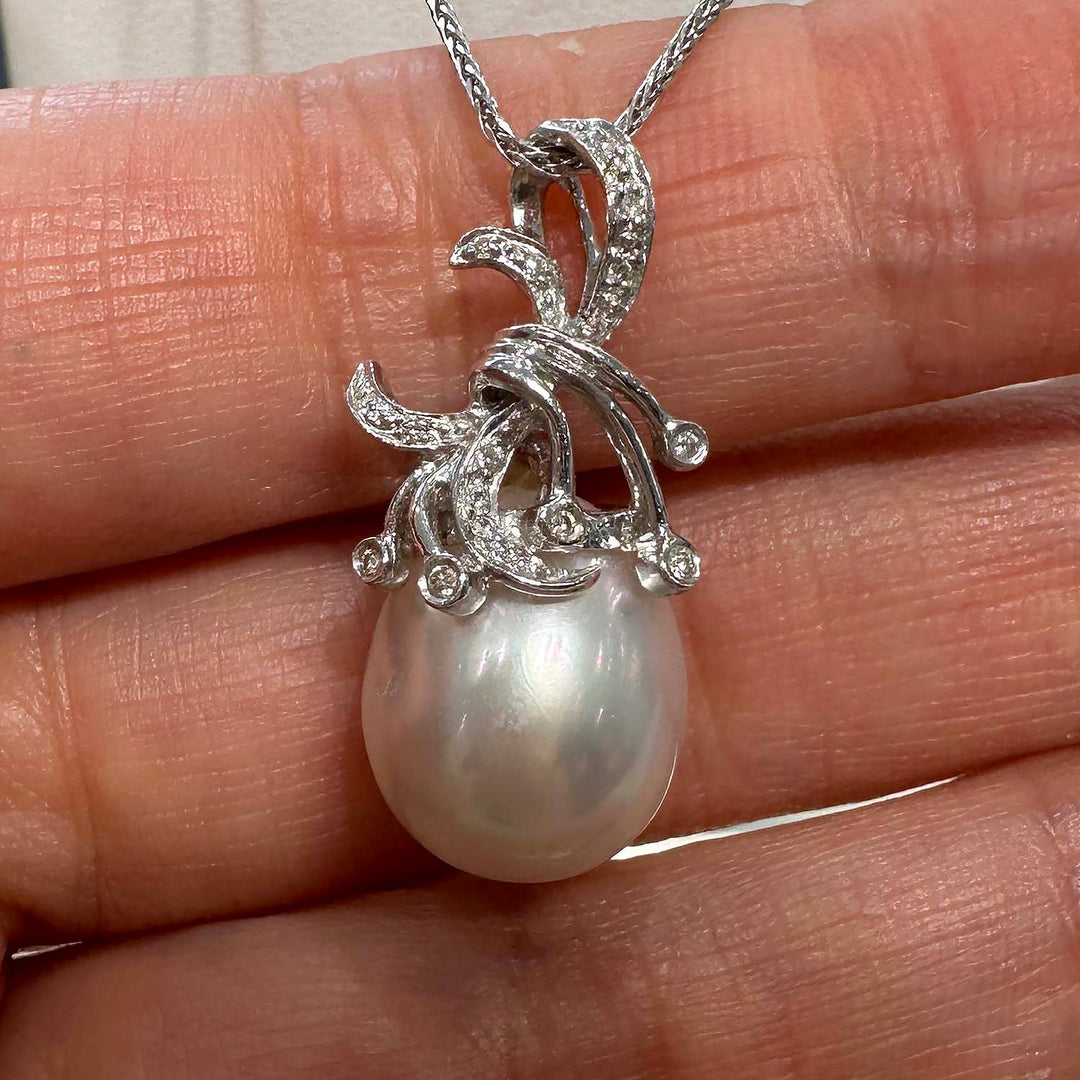 A close-up image of a pendant featuring a large South Sea pearl, suspended from an ornate silver setting adorned with small gemstones. The setting is designed with elegant, swirling motifs that add a decorative flourish around the pearl, which displays a lustrous sheen with subtle iridescent colors. The pendant hangs from a fine silver chain.
