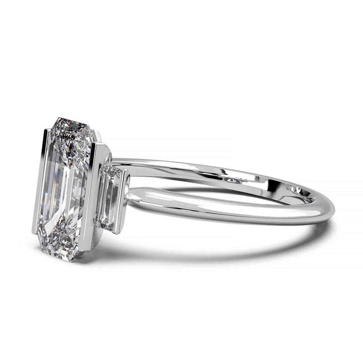 Three-stone engagement ring with emerald-cut diamond, baguette side stones, and modern bar prong setting.